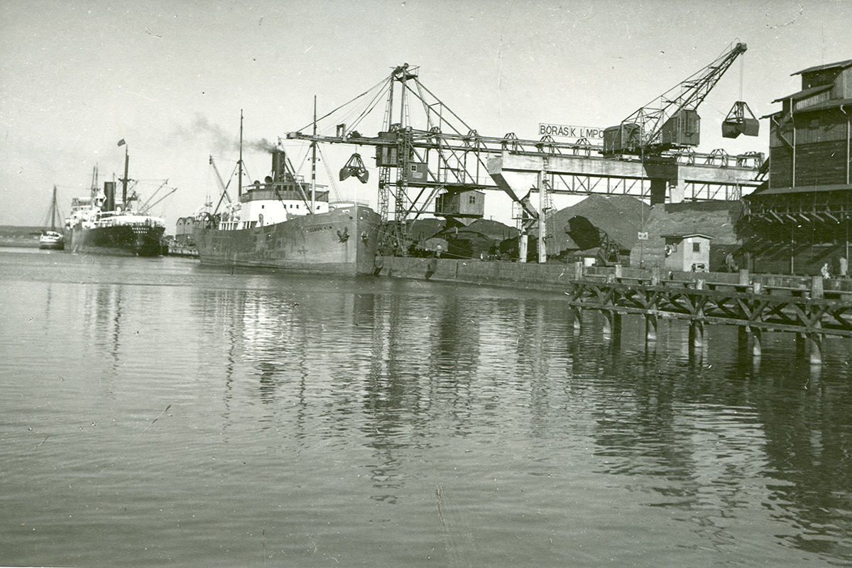 Gammalt foto från Varbergs hamn som visar kolkajen. Stora högar med kol ligger på kajen runt en stor kran.