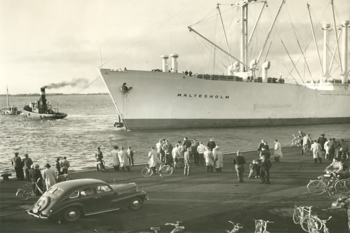 Varbergs hamn på 50-talet. Män i rock och hatt står på kajen och tittar på skeppet Maltesholm som lämnat Varberg med hjälp av lotsbåt.