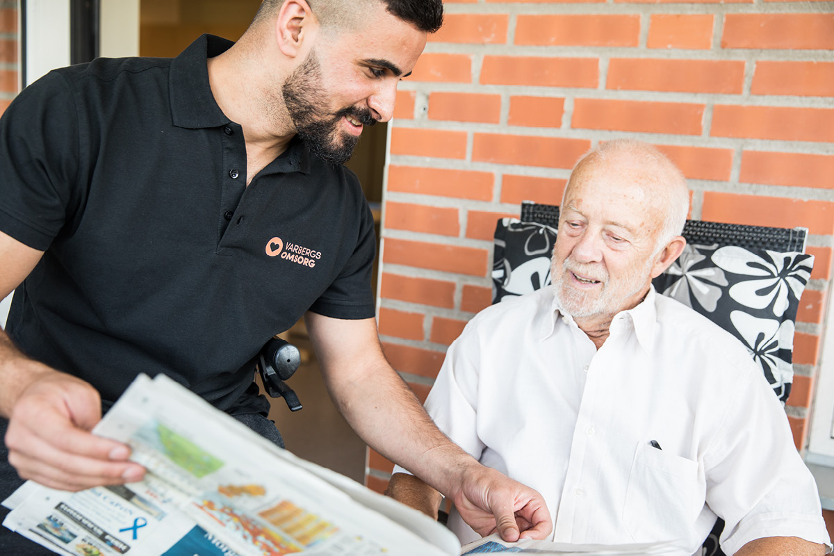 En manlig personal läser tidningen för en äldre man på ett äldreboende.