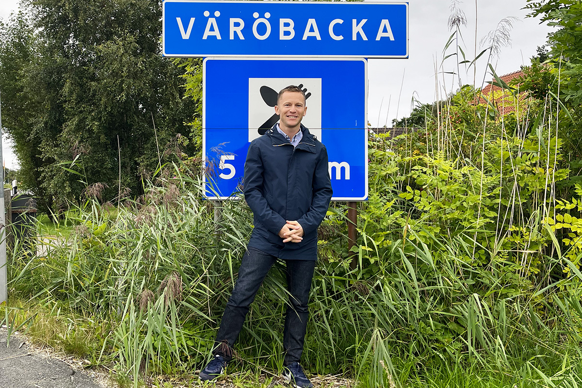 Man i 40-årsåldern står i en grön vägren med ryggen mot en vägskylt. På vägskylten står det Väröbacka med vita bokstäver på blå botten.