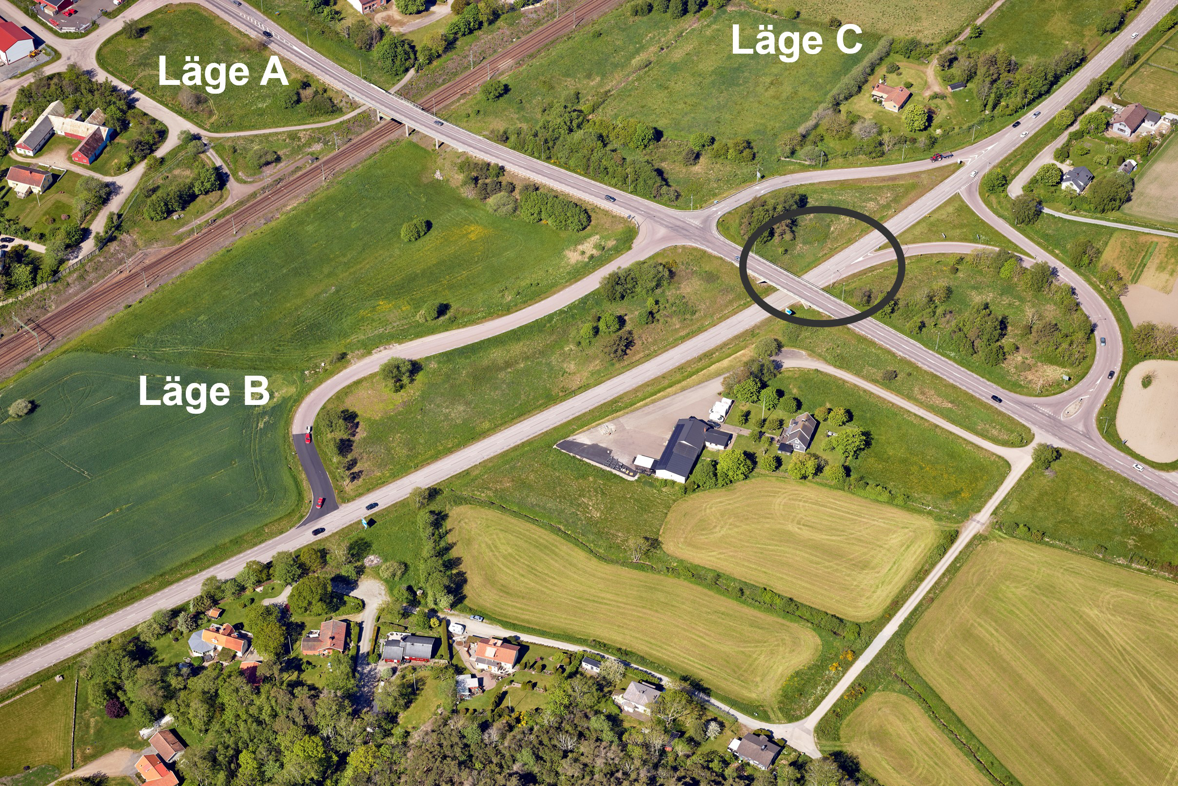 Flygbild över en trafikplats, mitt i ett jordbrukslandskap. I mitten en ritad rund svart cirkel som visar var en framtida rondell ska ligga.