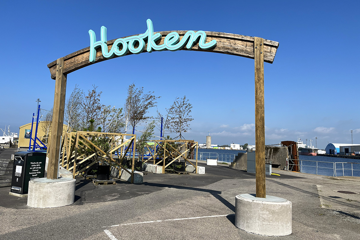 En stor skylt i trä med ljusblå neontext på tvärslån. Texten är ordet "Hooken". I bakgrunden skymtar en kaj och vatten.