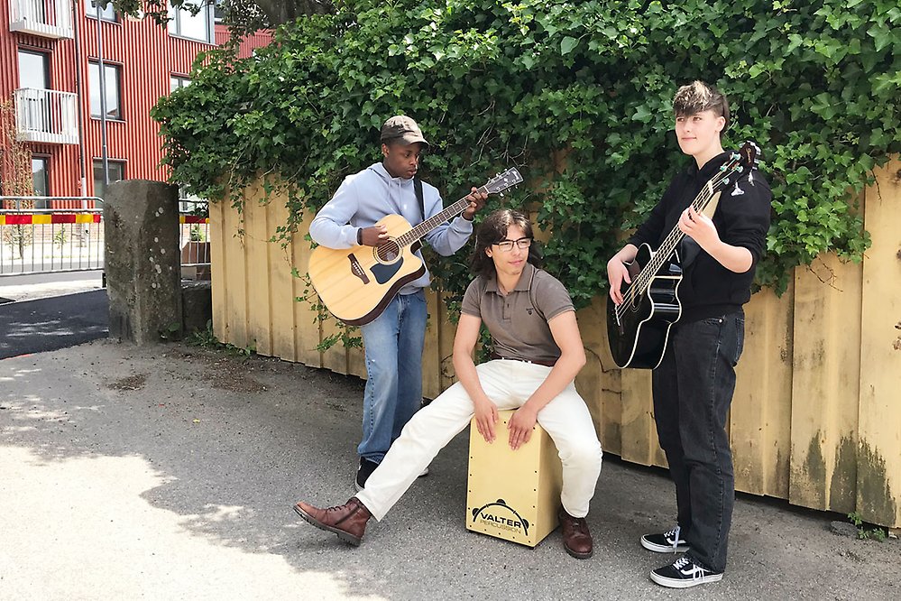 Tre unga personer står och spelar instrument i gatumiljö.